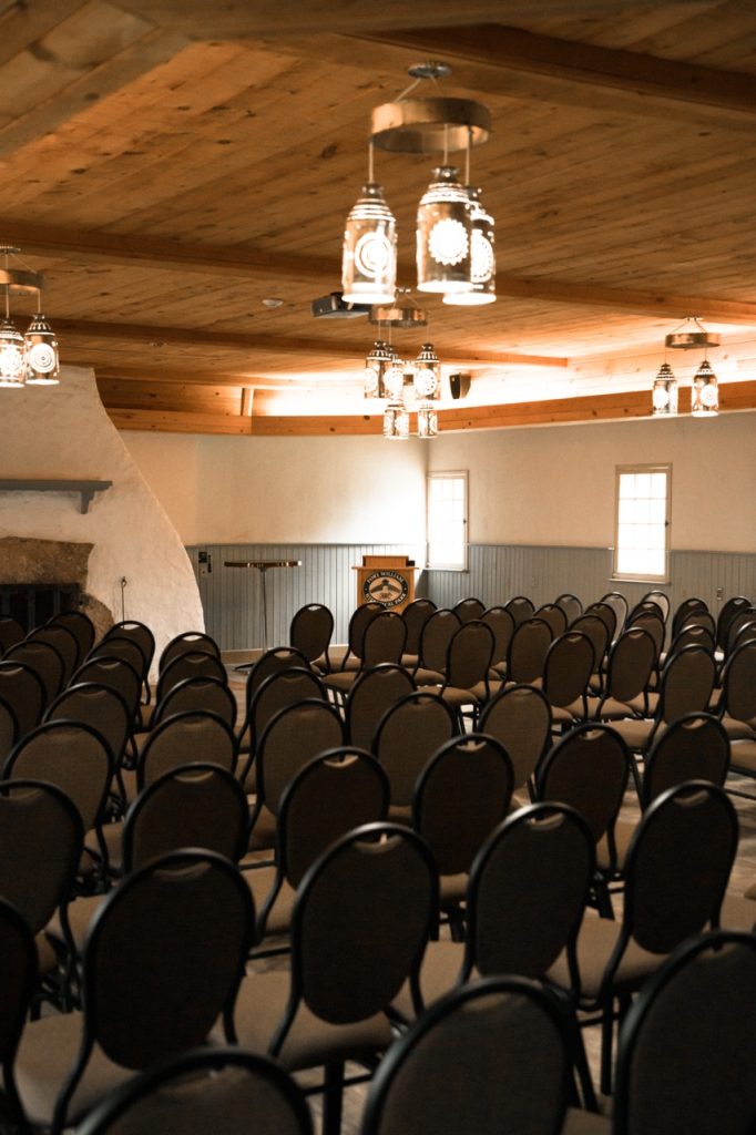 A side angle view of the Canot du Nord (North Canoe Shed) set up for a meeting with a center aisle and chairs in rows on either side facing a fireplace and podium