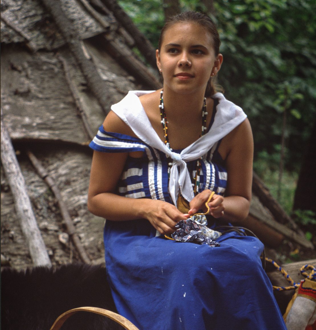A young girl dressed in historical dress sitting outside of a wigwam